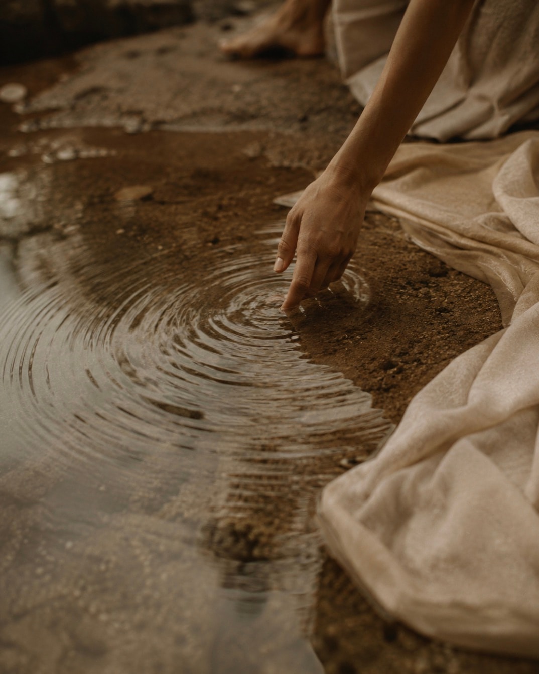 A hand touching shallow water beside softly draped natural fabric, creating gentle circular ripples.