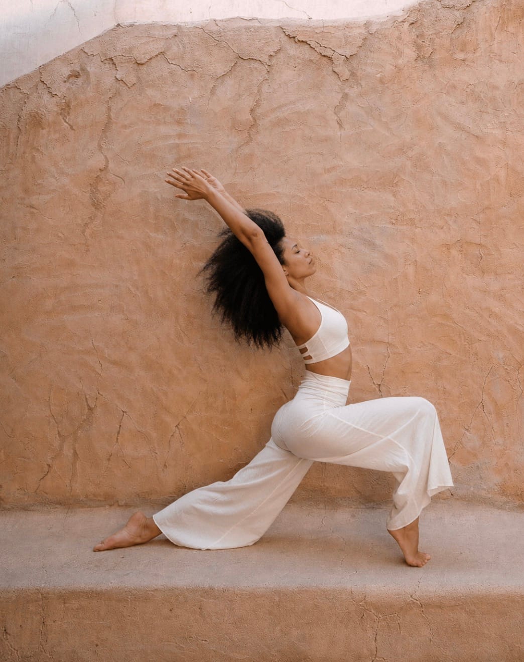 A woman in a flowing lunge stretch against a sunlit plaster wall, arm raised overhead, with generous space around her.