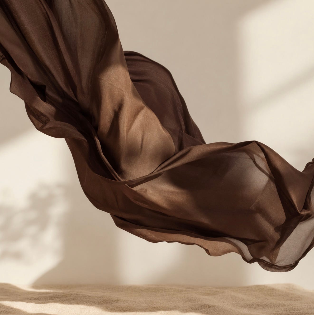 A sheer chocolate-brown fabric caught mid-motion, its folds warmly lit against a plain sand backdrop.