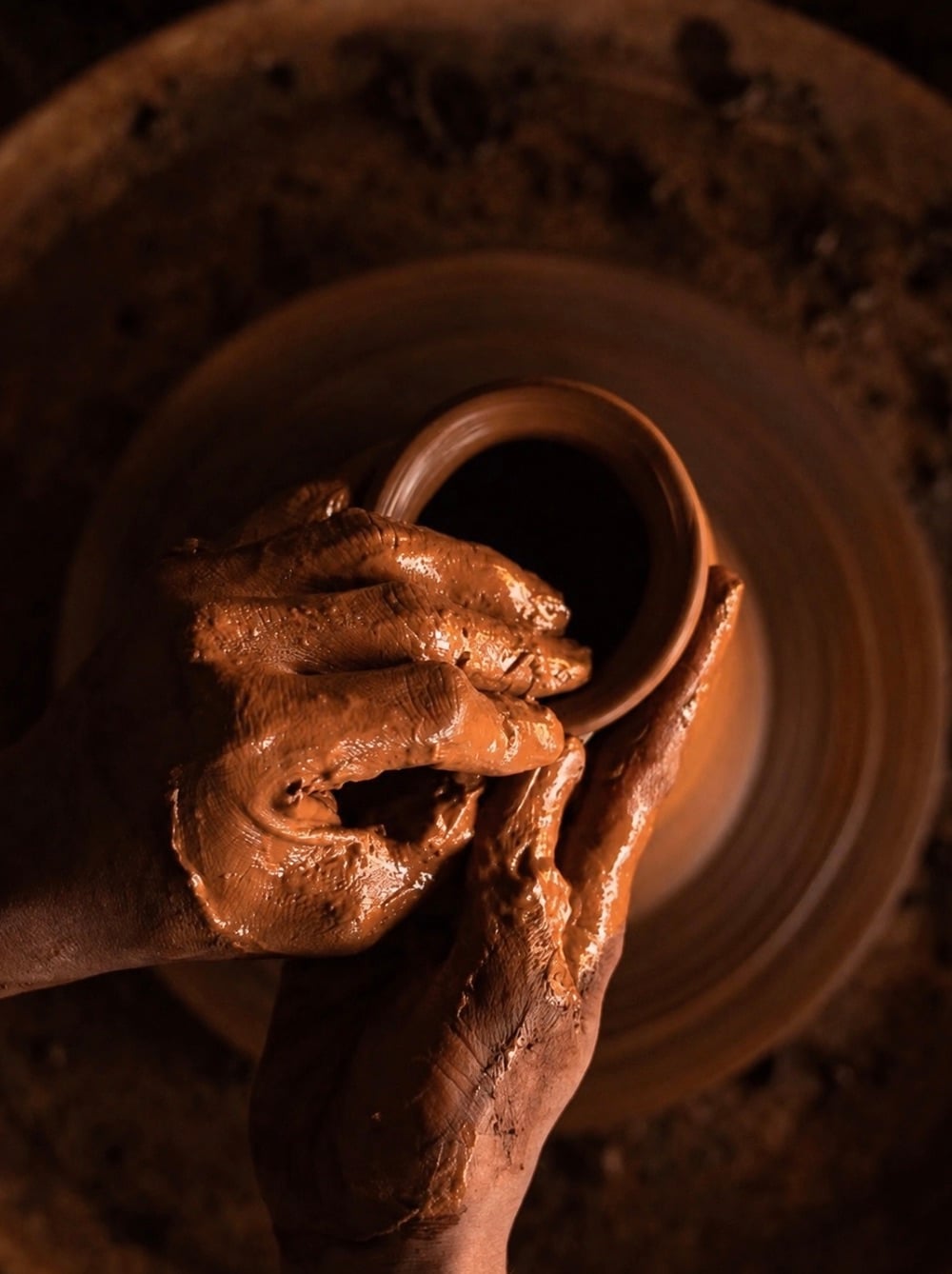Clay-covered hands shaping a vessel on a pottery wheel, seen from above.